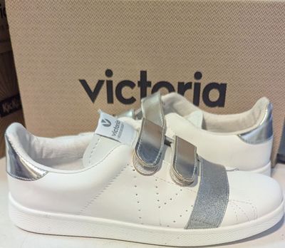 des baskets blanches et argentées, ornées de lanières argentées, exposées devant une boîte