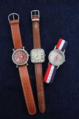 Trois montres sont posées sur une surface bleue, avec un bracelet rouge, blanc et bleu.