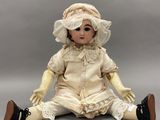 une poupée en porcelaine ancienne, assise par terre, vêtue d'une robe blanche et d'un bonnet