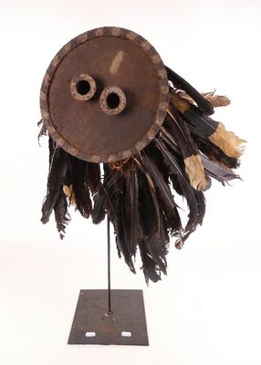 un masque marron orné de plumes, posé sur un socle, sur fond blanc