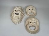 trois masques en céramique blanche représentant des visages, posés sur une surface blanche