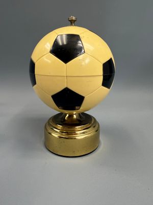 un ballon de football jaune et noir posé sur un socle doré, sur fond gris