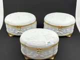 trois boîtes en porcelaine blanche, ornées de bordures et de poignées dorées, posées sur un fond noir