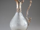un pichet ancien en argent et en verre posé sur un socle, sur fond gris