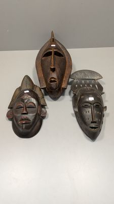 trois masques en bois posés sur une surface blanche, dont l'un porte un casque
