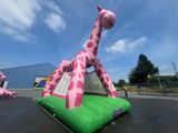 une structure gonflable en forme de girafe, rose et verte, installée sur un parking