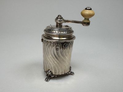 un moulin à café ancien en argent, muni d'un manche en bois, posé sur une surface blanche
